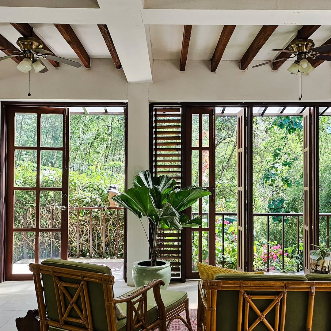 The heritage living room interior — rattan furniture, wood beam ceiling, and open louver windows looking out to the garden